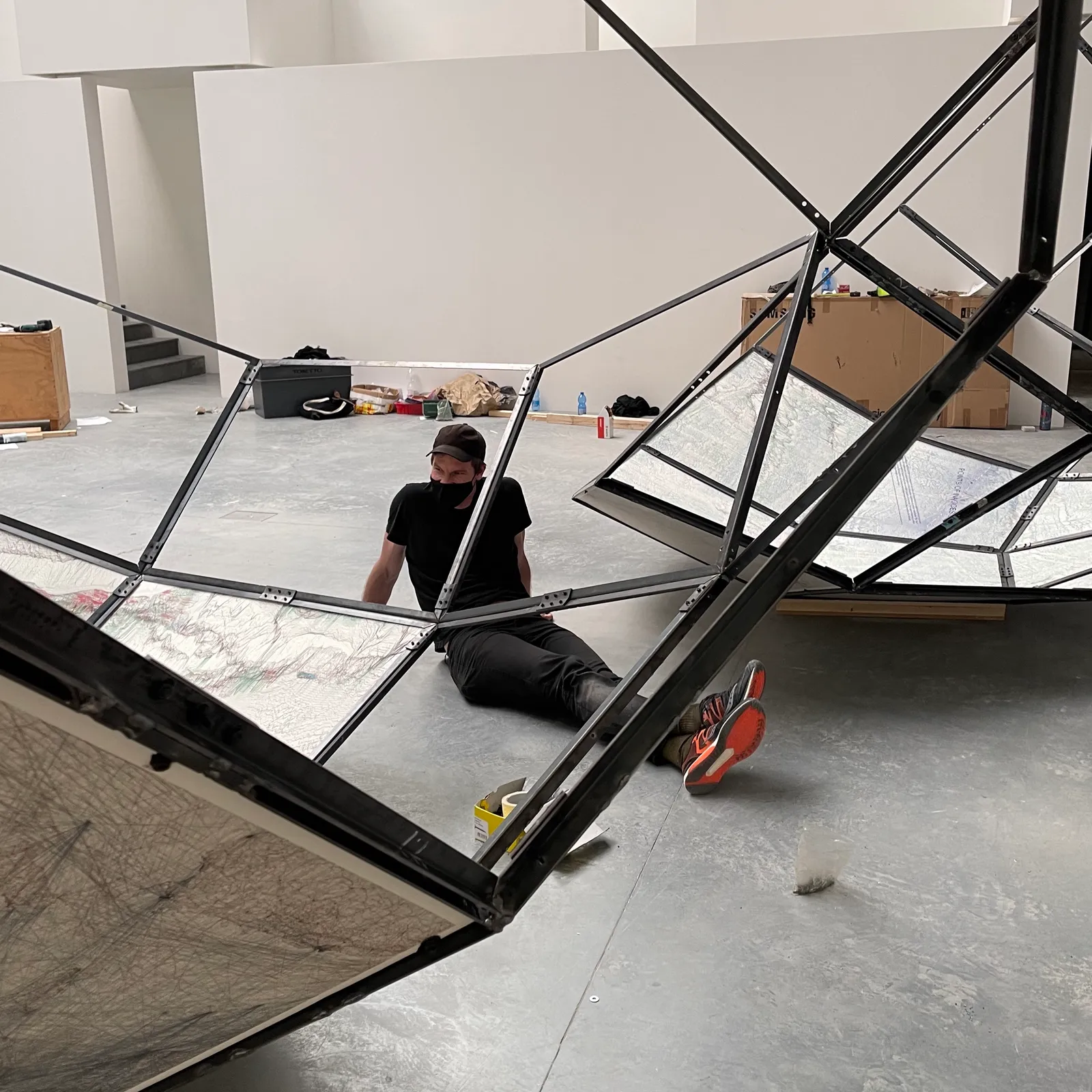 Jack Rusk, in a black cap and face mask, sits on the gallery floor inside the steel frame of a faceted map installation he is assembling — installing 'The World Turned Inside Out' at the Giardini for the 2021 Venice Biennale