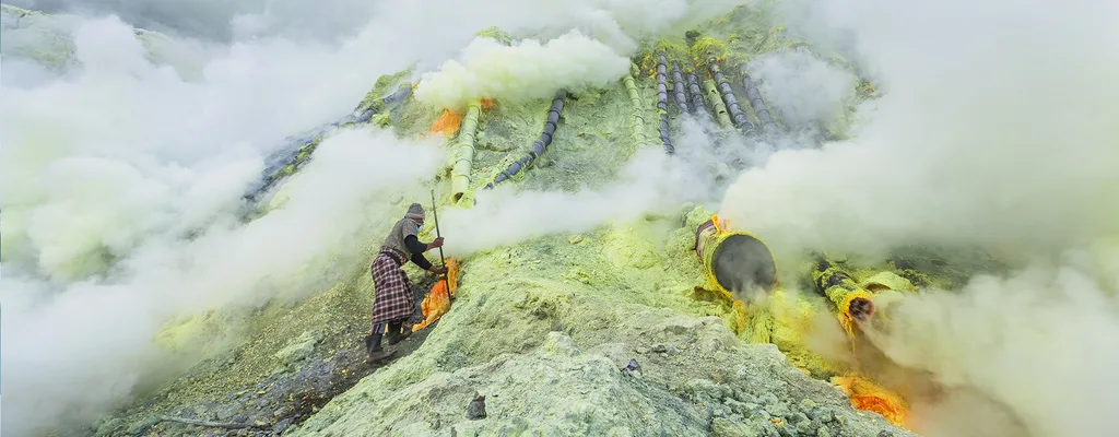 A sulfur miner in a plaid wrap working among smoking pipes and yellow sulfur deposits, half hidden in white fumes — header image for Jack Rusk's 2019 Commune essay 'A Tale of Two Parises'