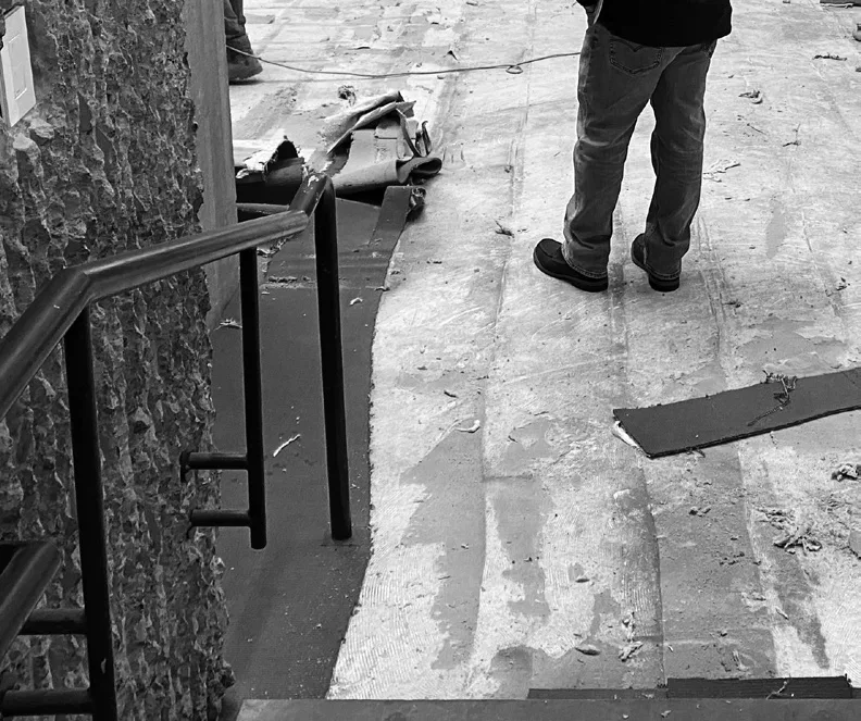 Black-and-white photo of a stair rail beside a rough concrete wall, with a worker standing on a floor stripped of its carpet — from Jack Rusk's 2020 Paprika! piece 'In Spite of the New Austerity'