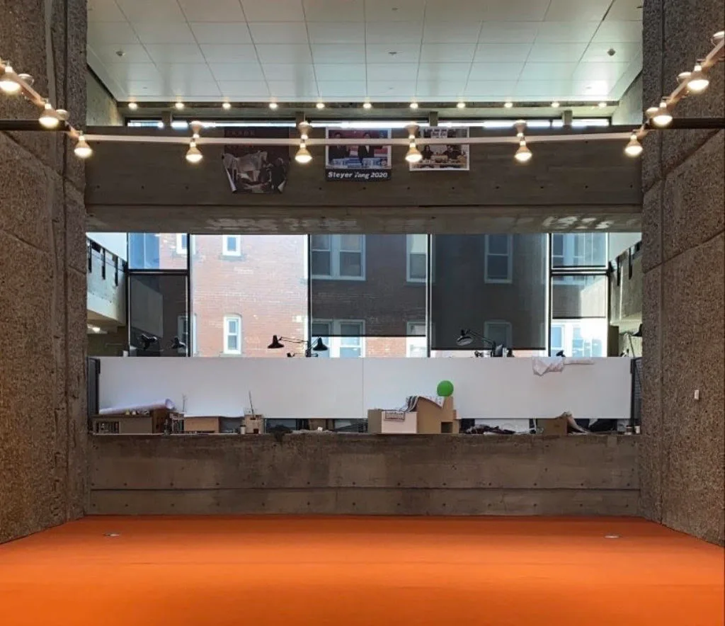 Photo of a gallery space with a bright orange carpet below a concrete balcony and studio windows — from Jack Rusk's 2020 Paprika! piece 'In Spite of the New Austerity'